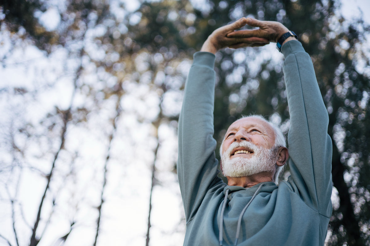 A man in his seventies stretching outdoors with his arms raised, smiling.